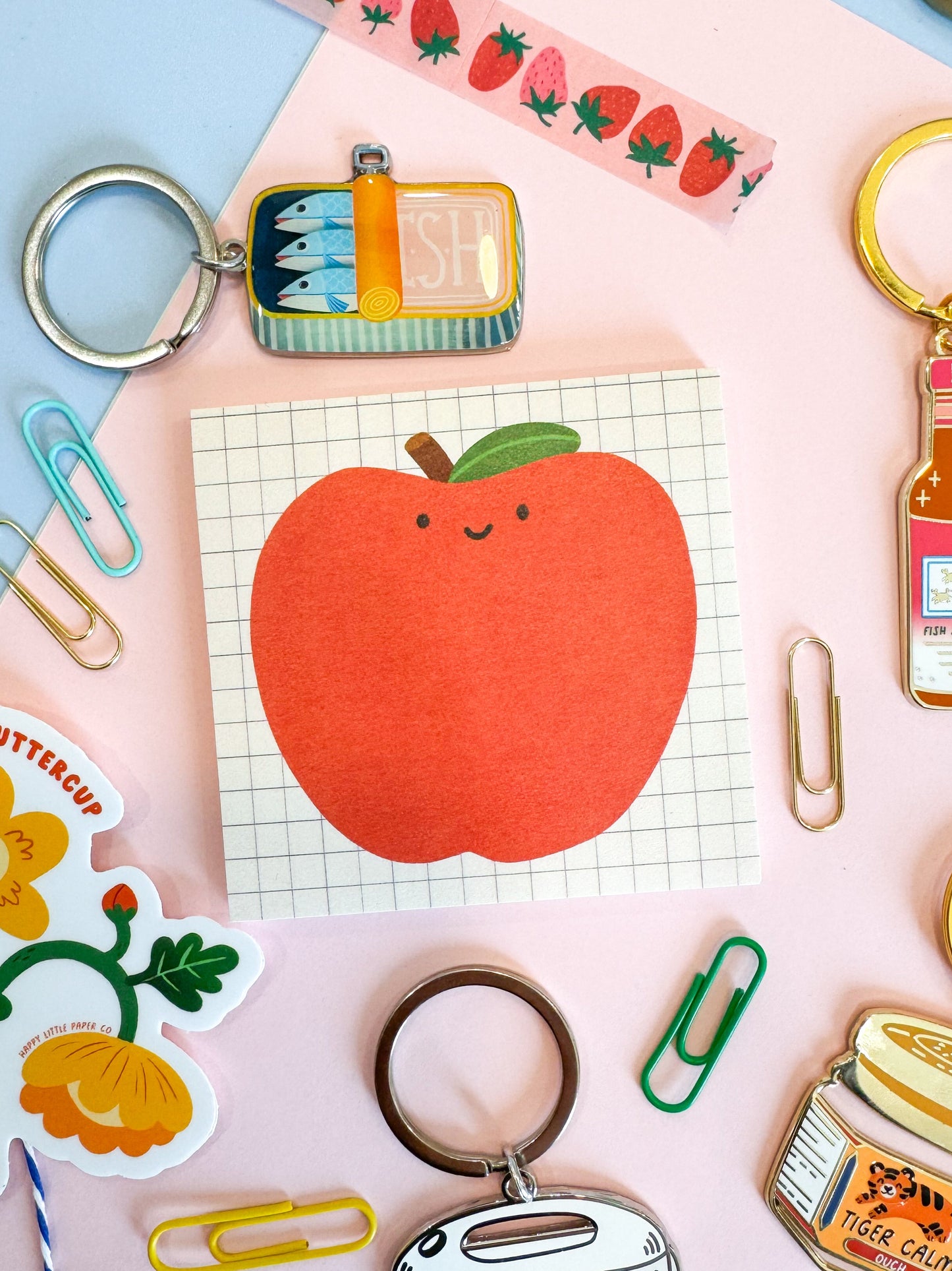 A styled flat-lay of multiple enamel keychains on a pastel blue and pink background. The keychains include a strawberry-patterned washi tape roll, a mini sandwich keychain, a hot sauce keychain, a jar of cream cheese keychain, a rice cooker keychain, and a sticky note with an illustrated apple on a grid pattern background.